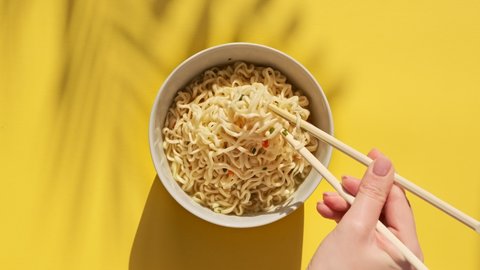 Instant noodles are mixed with chinese chopsticks on yellow background. Female hand mixes a hot meal. Top view egg noodles with spices in plate bowl.