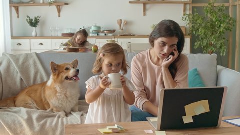 A Busy Mother Trying to Work Remotely With Her Children's at Home. The Daughter Interferes With Her Mother's Work. Working From Home During Quarantine Epidemic. Near Lying on the Couch a Cute Dog.