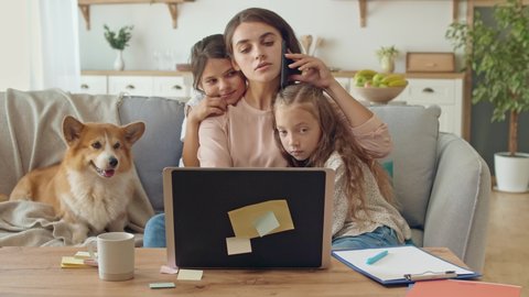 Lovely Mother Working Remotely From Home While Hugging Kids. Mother is Stroking a Daughters.A Busy Mother Working Remotely With a Laptop and Doing Business Conversation on the Phone Sitting on a Couch