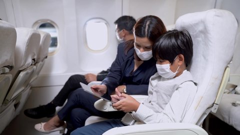 Interior of airplane with family and kid passengers with face mask sitting and using smart tablet device before take off. Concept family vacation in safety health for prevent pandemic disease.