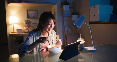 asian woman is eating instant noodles and tapioca ball milk tea while she watching video on digital tablet at home in the evening