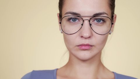 Close-up portrait of beautiful young woman looking at herself in the mirror and trying on a suitable frame of glasses on beige background. Optics selection concept
