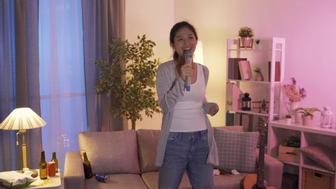 asian female standing dancing and waving hands is having fun singing karaoke in the evening in the living room surrounded by beer cans and bottles.