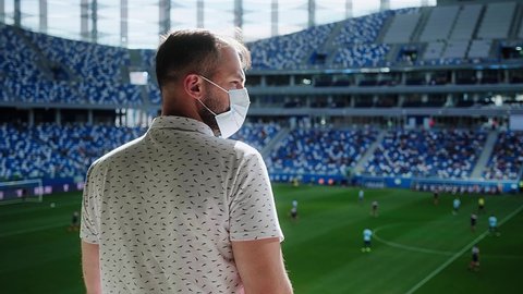 Стоковое видео: Young man in a summer sunny day in a medical mask watching a football match at the stadium. Mass event during coronavirus quarantine. High quality 4k footage