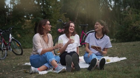 Three young cheerful women sitting on a blanket in the park drinking wine laughing and looking at the smartphone. : vidéo de stock