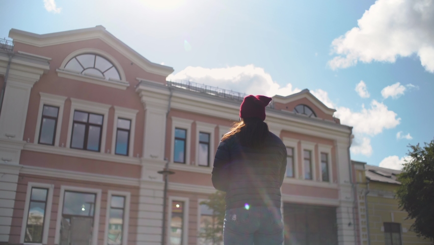 Back view, in a dark red hat, blue jacket and jeans, looks at the building, looks down, redraws, makes a sketch, layout, creates a picture, prepares a project, street, sunny weather, spring, autumn
