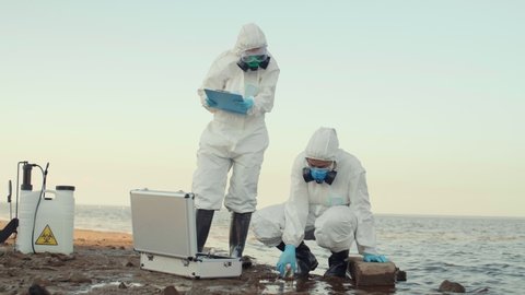 Wide shot of two scientist wearing protective coverall, goggles and masks taking water sample of polluted sea