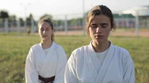 Two young girls in white kimonos sit on the grass and meditate with their eyes closed before karate training. Front view. Close-up. Blurred background