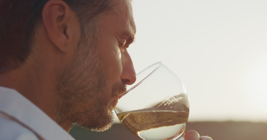 Authentic shot of happy handsome elegantly dressed man is tasting a flavor and drinking fresh white wine poured in transparent glass on scenic vineyards background at sunset.