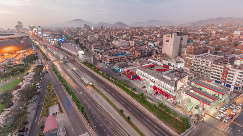 Aerial top view of Via Expresa highway and metropolitan bus with traffic day to night transition timelapse near National Stadium of Peru. Illuminated houses and city skyline with mountains