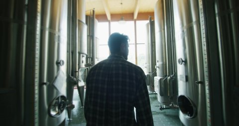 A happy successful farmer or winemaker is walking in the middle of wine tanks and checking a fermentation grade of high quality wine production in a modern winemaking facility or winery.