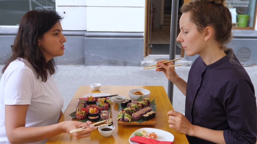 Brunette girl feeds her blonde friend sushi roll. Sharing food and good times with friends in the city | Shutterstock HD Video #1059714122