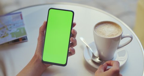 Crop view of female person holding smartphone and pressing on mockup screen while sitting at table with coffee and map on it. Concept of greenscreen and chroma key