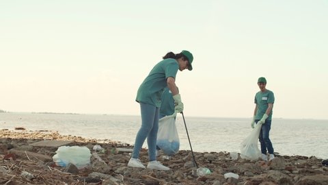Wide shot of group of three young volunteers picking up garbage on polluted bank of river : vidéo de stock