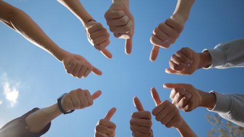 Bottom view of young people standing in a circle and showing thumbs up. Students or business team gesturing thumbs up over blue sky background outdoors
