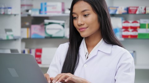 Doctor woman working in pharmacy store at hospital. Asian pharmacist girl looking laptop computer checking medication details in pharmacy drugstore. Concept of medical technology, business, lifestyle.