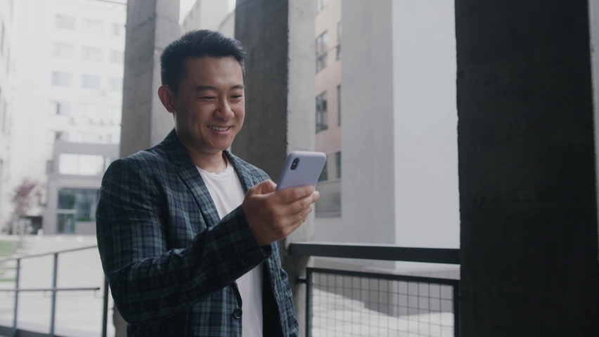 Happy asian young entrepreneur browsing mobile phone internet during office break walking along business district. Successful businessman going to work. Royalty-Free Stock Footage #1059722369