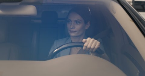 View through windscreen. Beautiful smiled Caucasian young businesswoman sitting at steering wheel, wearing seat belt and driving expensive car. Attractive cheerful female driver turning.