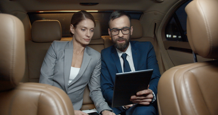Caucasian businessman and businesswoman sitting on backseat of car and talking on videochat via tablet device webcam. Couple of business partners smiling and videochatting. Video call. Online chatting Royalty-Free Stock Footage #1059727541
