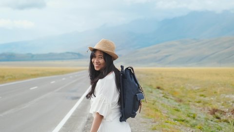 Woman having a trip in the mountains, she walks on the highway with backpack, turning back and smiling into camera. Backpacker travels and discovers world. Happy hipster asian traveller. Stock Video