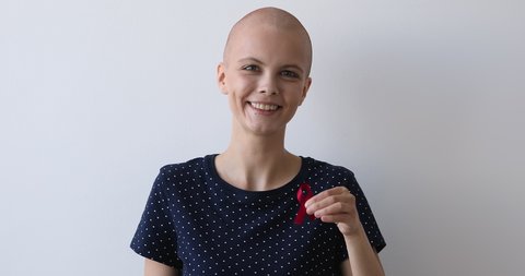 Head shot portrait bald young woman hold red ribbon pose on grey studio background. HIV AIDs awareness disease prevention protection, volunteering activity and charity. World breast cancer day concept