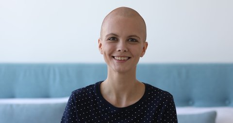 Head shot portrait pretty young bald woman sit on bed in bedroom alone smiling looks away feels hopefulness and confidence about the healthy future and success of innovative cancer treatments concept