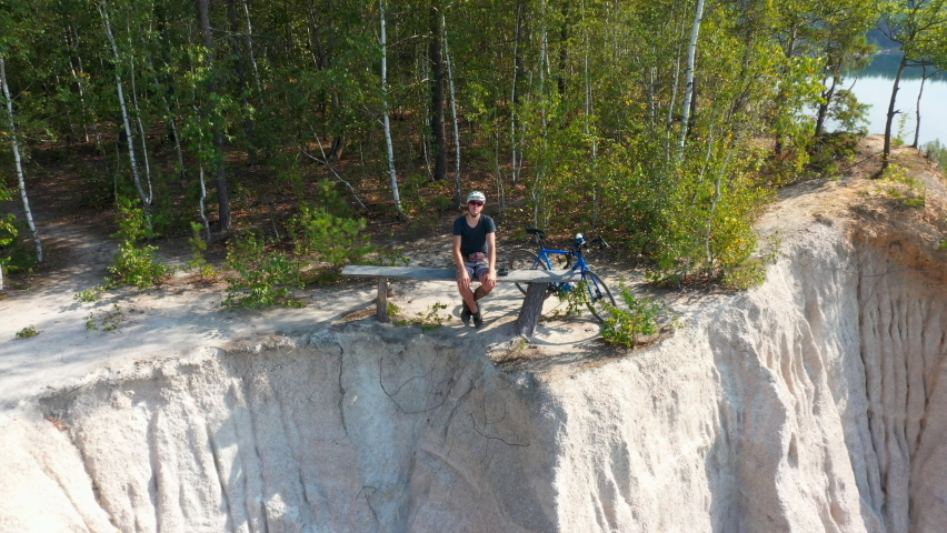 Man with  bicycle sitting on a bench near a mountain of rubble near a lake in the forest. Drone view of the lake in the forest. Aerial view.