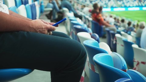 A man sits on the stadium stand and surfing social media on his smart phone during a football or soccer match. Mass event during coronavirus quarantine. Close-up of hands. High quality 4k footage
