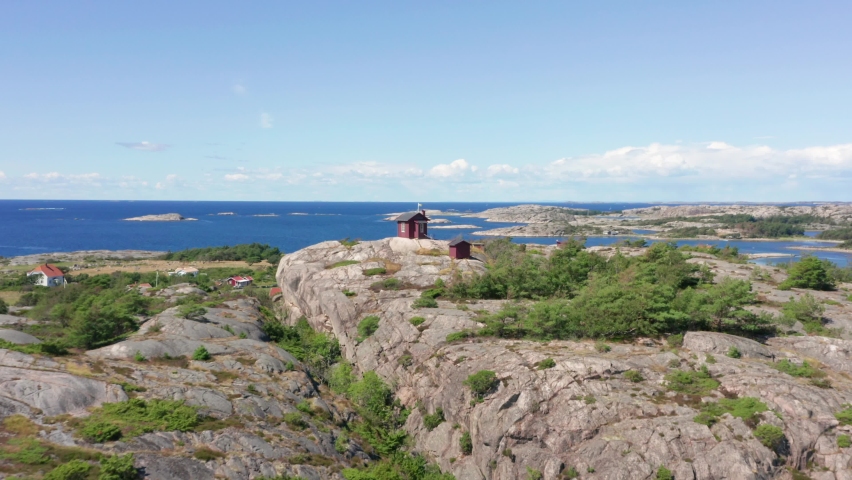 Coastline in Sweden. Aerial drone shot of tiny red summer house on mountain rock and granite rocky islands in background. Swedish archipelago on west coast Bohuslan. Flag waving in light summer wind Royalty-Free Stock Footage #1059741764