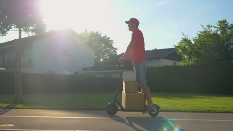 LENS FLARE: Young male courier delivers packages on his e-scooter on a sunny summer day. Cinematic shot of a millennial parcel delivery guy having fun transporting packages on his electric scooter.
