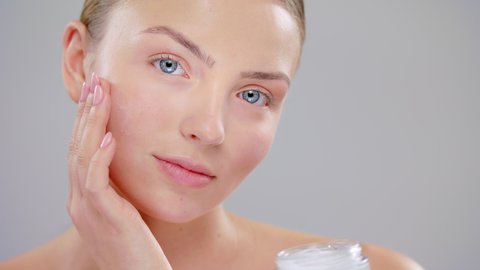 Close Up Beautiful Young Caucasian Blonde Woman With Blue Eyes And Smooth Healthy Skin Holding A Container Of Cream And Using White Cream Skin Care Cosmetic Concept Slow Motion On Grey Background
