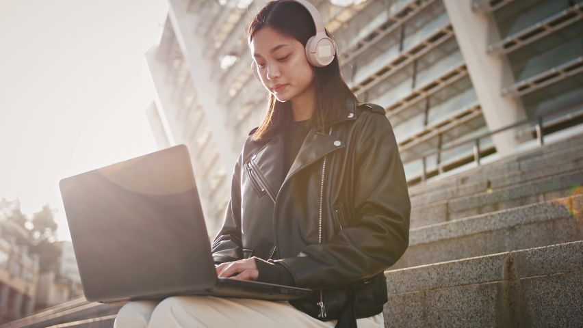 Asian female freelancer in headphones is working online, typing on notebook while sitting on stairs near a stadium