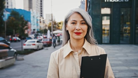Asian mature business woman is smiling, holding black folder, looking at you while posing on street of city with busy traffic
