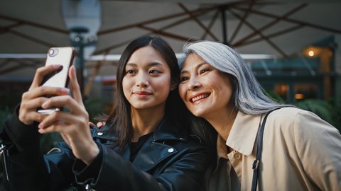 Asian middle-aged mother and adult daughter taking selfie by smartphone and smiling while posing near terrace of cafe