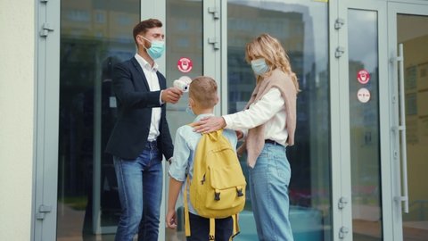Security guard at school checking temperature of young junior student. Beautiful mom accompanies her son to the school building. Face masks. Virus prevention.
