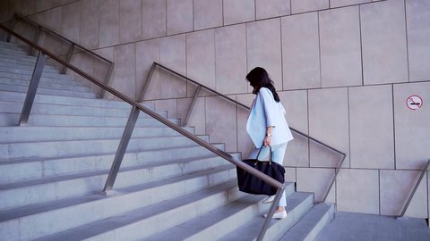 Beautiful attractive business woman hurries looks at watch and walking up stairs to office building. Busy brunette running to work with laptop and big black bag, wear blue suit.