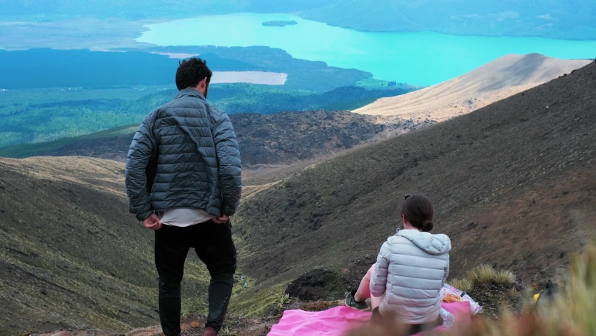 Young couple picnicking high in the mountains and looking over a valley of hills, green fields and a lake - over which shadows of clouds moving - Panning Time-Lapse