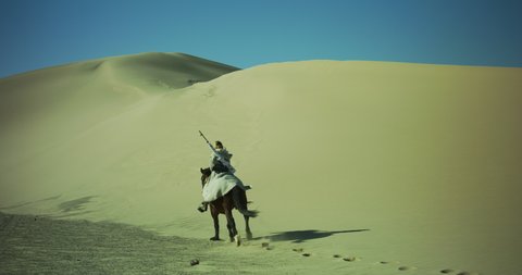 Masked person rides a galloping horse through the desert up a sand dune. Sand dunes surround a person riding a horse with a rifle on their back. Long journey. Cinematic, Shot on RED