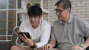 Happy two age generations asian men family old father embracing young adult son having fun enjoying using digital tablet watching funny social media video using mobile apps at home sit on sofa.
