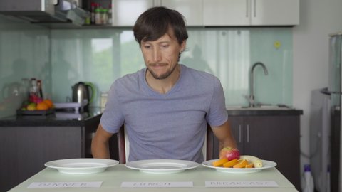 A sad young man puts signs fasting on empty plates with signs lunch and dinner and then eats an apple from the breakfast plate. Interval fasting concept. Skipping meals
