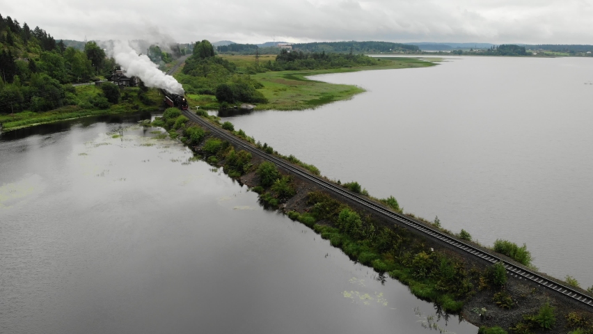 Historic Old steam locomotive train with a wagons rides going along the embankment of Lake. there is black grey smoke. Russia, Karelia, Sortavala. Flying over. Aerial view. Drone is standing Royalty-Free Stock Footage #1059768167
