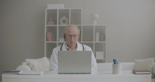 aged male physician is working remotely from his office in clinic, using laptop for communication with colleagues