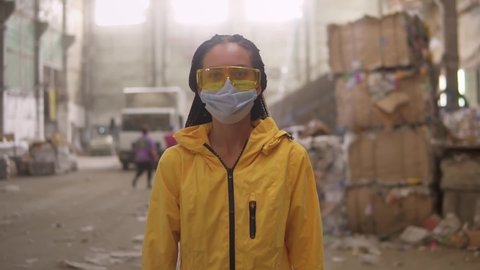 A positive girl in protective working clothes - yellow jacket, eyeglasses and mask walking by recycling factory and smiling. Boxes of waste, truck on background