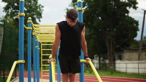 Muscular, bearded man doing push ups on crossbar outdoors on stadium in summer