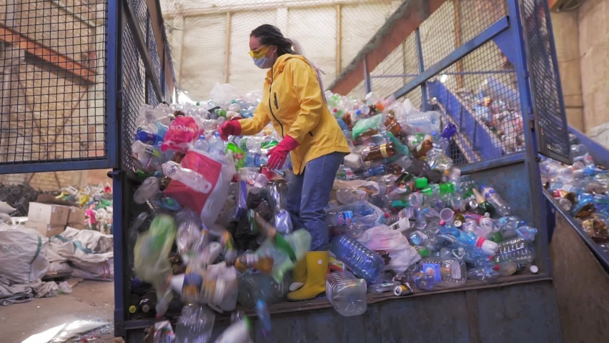 Footage of a young woman in yellow jacket and gloves scoops used bottles by hands used plastic bottles at recycling factory. Huge pile of bottles inside the car case cage Royalty-Free Stock Footage #1059771728