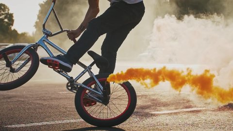 Young male is performing difficult tricks riding bmx bike with orange smoke bomb attached to its rear wheel