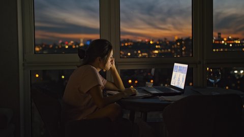 Remote work. Young woman networking on laptop, sitting at window with great cityview late in evening, side view Adlı Stok Video