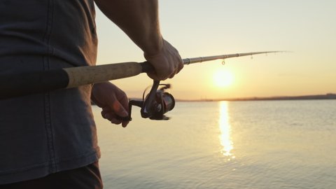 Handle rotation with reel of fishing rod against of orange sunset slow motion. Man hobby fishing on sea tightens a fishing line reel of fish summer. Lens flare. Calm surface sea. Bright disk of sun