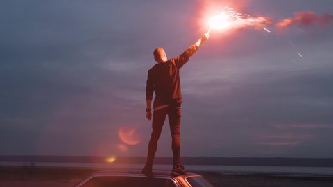 Стоковое видео: Handheld cinematic dark shot of young man standing on the car with red signal burning flare on the beach near the water