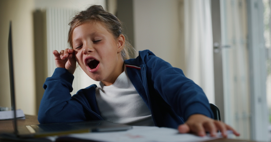 Authentic shot of little girl pupil is yawning tired while sitting at desk and writing in notebook while doing a homework to prepare for school day in her room at home.